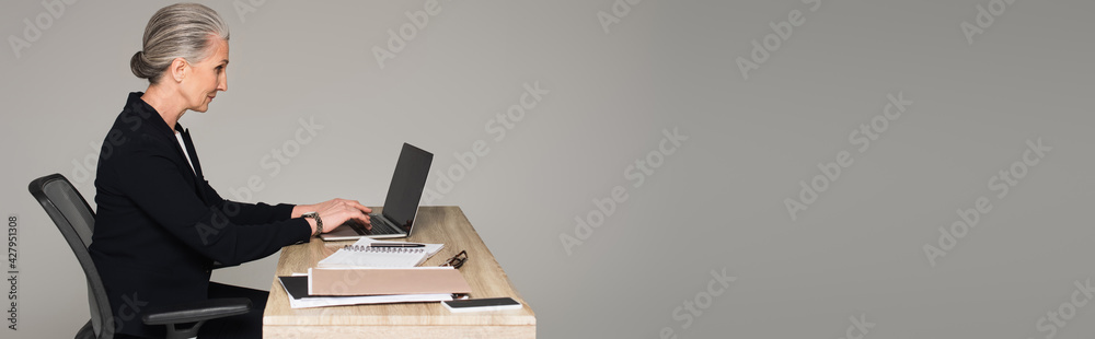 Side view of mature businesswoman using laptop near gadgets isolated on grey, banner Side view of mature businesswoman using laptop near gadgets isolated on grey, banner