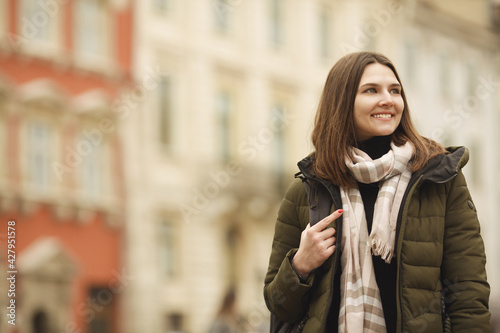 Travel concept: happy smiling young woman posing at street of European city. Model wearing green coat, white and beige scarf. Copy, empty space for text. Outdoor shot © Augustino