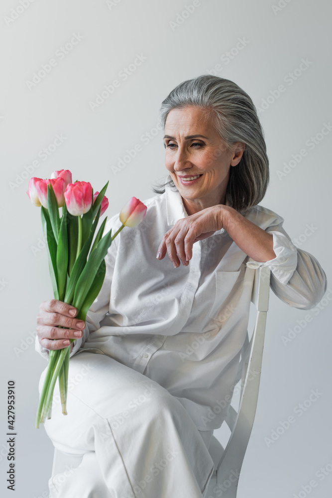 Grey haired woman in white shirt looking at flowers on chair isolated on grey Grey haired woman in white shirt looking at flowers on chair isolated on grey