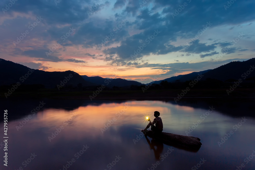 Beautiful landscape Silhouette image with mountains reflecting the blue sky of the river. The clouds and the orange light in the sunset and the shadow of a man with a mobile flash.