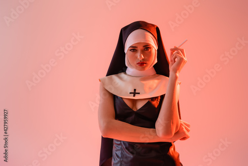 Fotografiet sexy nun in leather dress looking at camera while holding cigarette isolated on