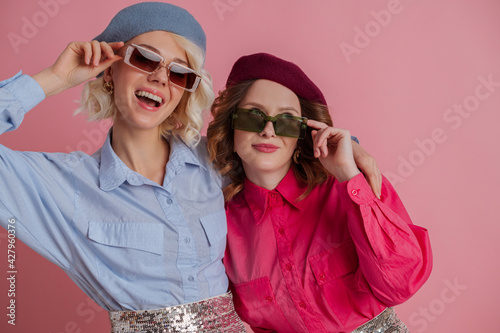 Two happy smiling girls wearing stylish sunglasses, colorful clothes posing on pink background. Copy, empty space for text