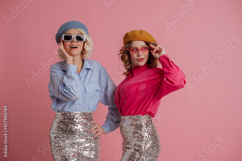 Spring fashion, advertising concept: two happy smiling women, friends, sisters, wearing trendy sunglasses, colorful shirts, berets, posing on pink background. Copy, empty space for text