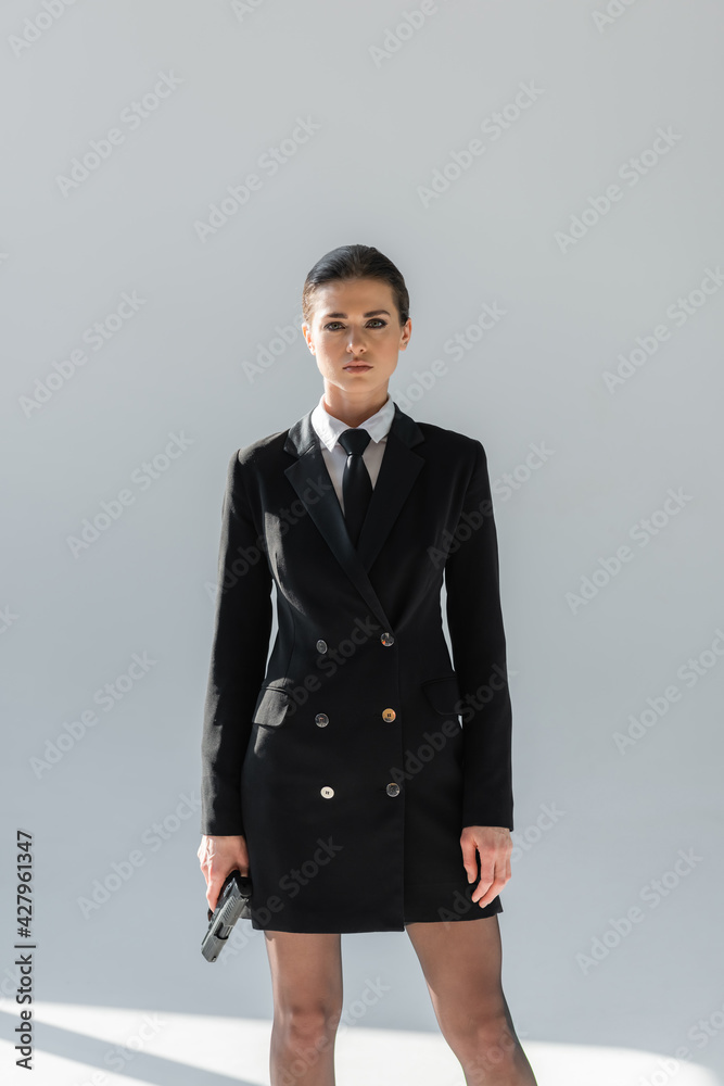 elegant woman in black blazer dress looking at camera while holding gun on grey background elegant woman in black blazer dress looking at camera while holding gun on grey background