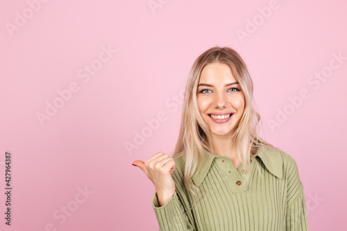 Pretty european woman in casual sweater on pink background happy smiling point left with thumb finger