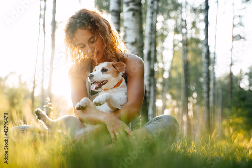 Young red-haired woman sitting on green grass in the park holding jack russell dog in her arms.