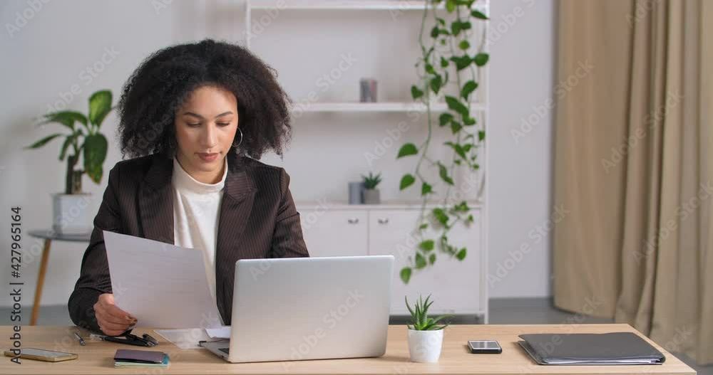 Afro american girl student secretary freelancer business woman sitting at table in home office studing looking at laptop reads reports compares data on paper and on computer screen analyzes project Afro american girl student secretary freelancer business woman sitting at table in home office studing looking at laptop reads reports compares data on paper and on computer screen analyzes project