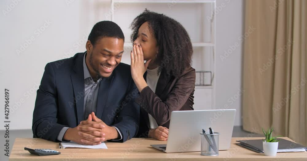 Afro american colleagues man and woman sitting in modern office in business company girl tells secret to male colleague share funny gossip joke good news confidential information man laughs rejoices Afro american colleagues man and woman sitting in modern office in business company girl tells secret to male colleague share funny gossip joke good news confidential information man laughs rejoices
