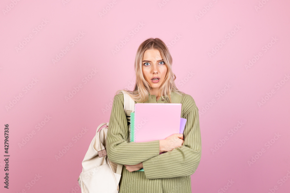 Pretty european woman in casual sweater on pink background with notebooks shocked look to camera with open mouth, education high school concept Pretty european woman in casual sweater on pink background with notebooks shocked look to camera with open mouth, education high school concept