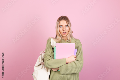 Pretty european woman in casual sweater on pink background with notebooks shocked look to camera with open mouth, education high school concept