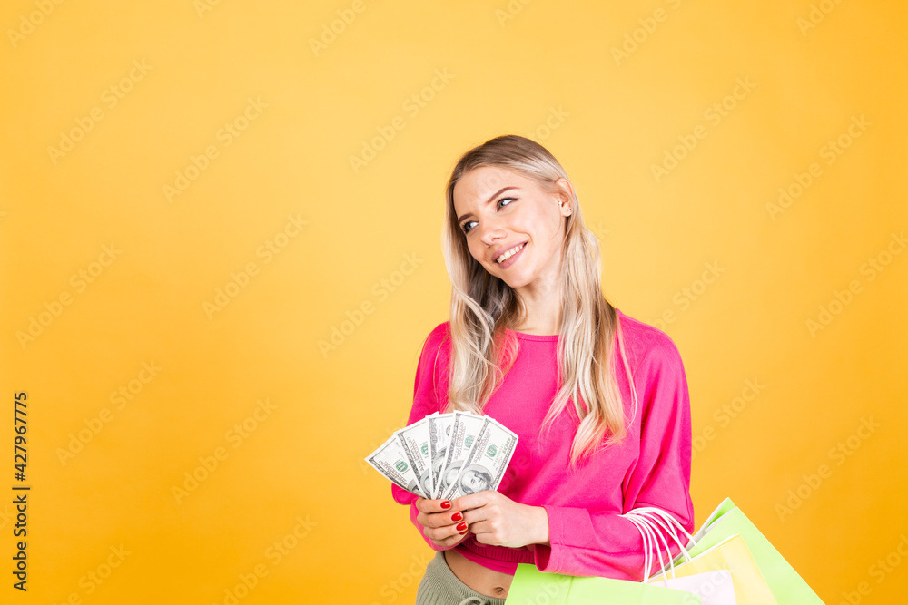 Pretty european woman in pink blouse on yellow background holding fan of 100 dollar bills money and shopping bags thinking positive smiling copy space Pretty european woman in pink blouse on yellow background holding fan of 100 dollar bills money and shopping bags thinking positive smiling copy space