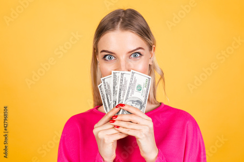 Pretty european woman in pink blouse on yellow background hold fan of 100 dollars money and, excited surprised cover face
