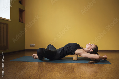 Woman in black sportswear practicing yoga doing Supta baddha konasana exercise with a wooden block under her back on a mat in the studio