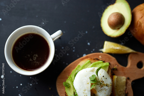 Cup of black coffee and sandwich with avocado and poached egg, seeds and microgreen. Black background. Copy space. Healthy breackfast