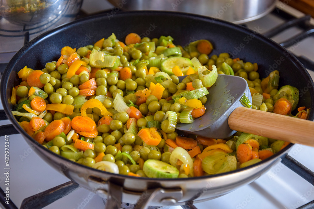 Process of cooking saute at home. Stewed vegetables and silicone spatula with wooden handle in frying pan. Mixed Ingredients leek, carrot, zucchini, bell pepper, green peas. Close-up. Selective focus. Process of cooking saute at home. Stewed vegetables and silicone spatula with wooden handle in frying pan. Mixed Ingredients leek, carrot, zucchini, bell pepper, green peas. Close-up. Selective focus.