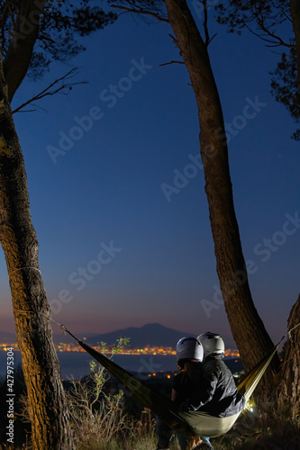 Couple relaxing in a hammock and enjoying starry sky. View from the top of the mountain to the lights of the city. Volcano Vesuvio. Sea and romance. Vertical photo. Motorcyclist. Helmet. Biker photo