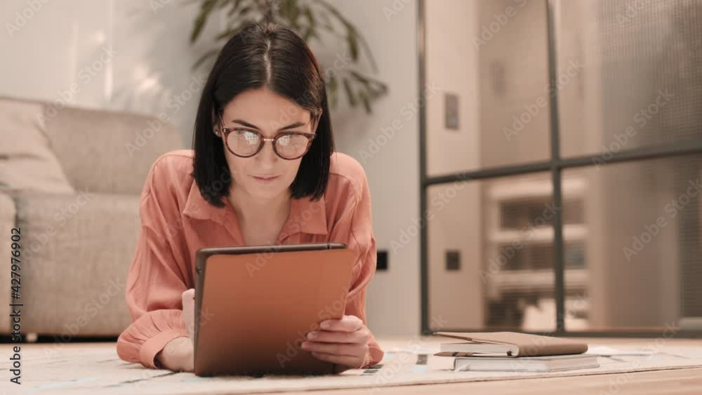 Medium close-up of young black-haired Caucasian woman wearing glasses, lying on stomach on floor, using tablet computer Medium close-up of young black-haired Caucasian woman wearing glasses, lying on stomach on floor, using tablet computer