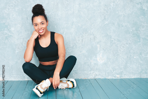 Fitness smiling black woman in sports clothing with afro curls hairstyle.She wearing sportswear. Young beautiful model with perfect tanned body.Female sitting in studio near gray wall
