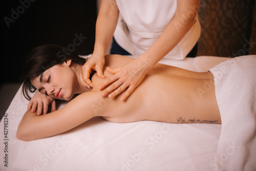 Beautiful naked girl with perfect skin with closed eyes is relaxing during back and shoulder massage lying on massage table in spa salon. Concept of luxury massage. Concept of body care.