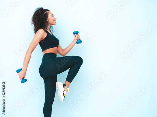 Fitness smiling black woman in sports clothing with afro curls hairstyle.She running and jogging . Young beautiful model with perfect tanned body.Female holding dumbbells in studio near blue wall