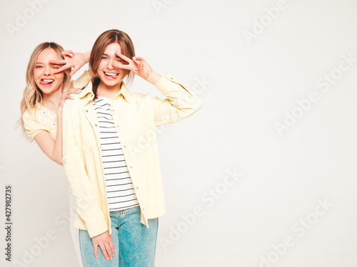 Two young beautiful blond smiling hipster female in trendy summer clothes. Sexy carefree women posing on grey background in studio. Trendy and positive models having fun.They show peace sign © halayalex
