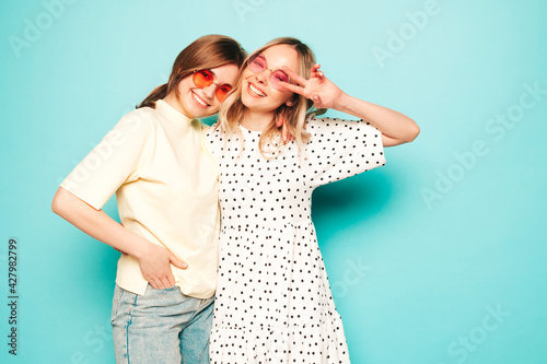 Two young beautiful blond smiling hipster female in trendy summer clothes. Sexy carefree women posing near blue wall in studio. Trendy and positive models having fun in sunglasses © halayalex