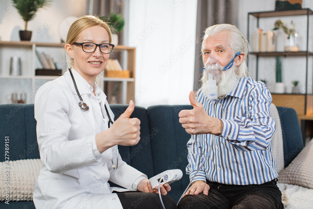 Senior grey-haired bearded man patient wearing nebulizer inhaler mask, sitting with his smiling professional family doctor, and looking at camera with thumbs up Senior grey-haired bearded man patient wearing nebulizer inhaler mask, sitting with his smiling professional family doctor, and looking at camera with thumbs up