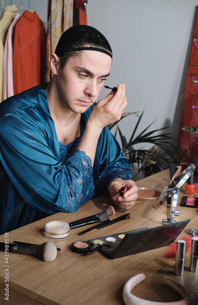 Obraz premium Young transgender man applying makeup at the table he applying mascara on his eyes