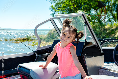 Happy little girl sits in a motor boat on the shore of a lake or river. Summer vacation. Rest with children by the water. 