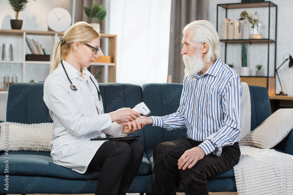 Obraz premium Middle aged practitioner visiting sick elderly man patient at home. Young blond woman doctor measuring patient temperature with non-contact wrist infrared thermometer at home