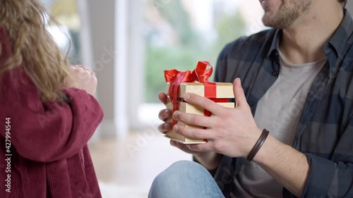 Unrecognizable daughter giving gift box to surprised father indoors. Little Caucasian girl and young man celebrating holiday at home. Family lifestyle photo