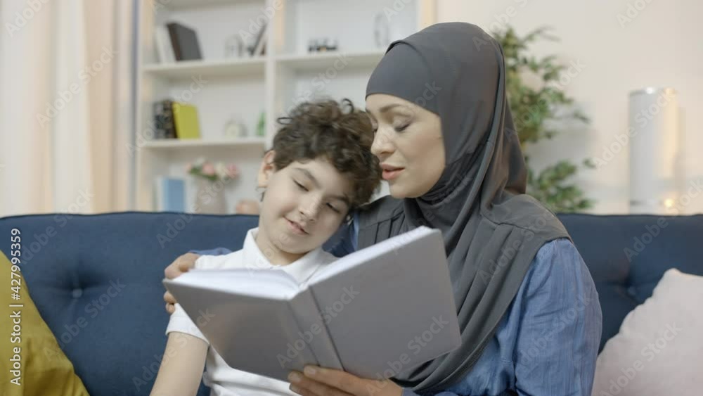 Caring muslim mother reading book to little son, enjoying time together, family