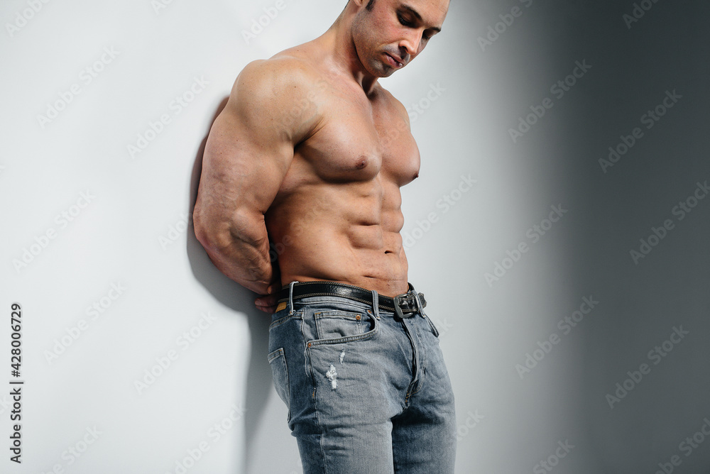 A young athlete bodybuilder poses in the studio topless in jeans near the wall. Sport. A young athlete bodybuilder poses in the studio topless in jeans near the wall. Sport.