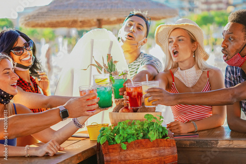 Multiethnic friends drinking coktails in a beach bar outside in summer days with face mask on to be protected from coronavirus -  Happy people cheering with mojito and having fun © BooNKer