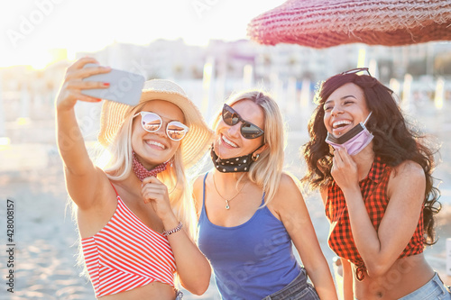 Teenagers taking a selfie in beach - Young girls smiling in front of the camera with face mask on - The new coronavirus lifestyle of this summer