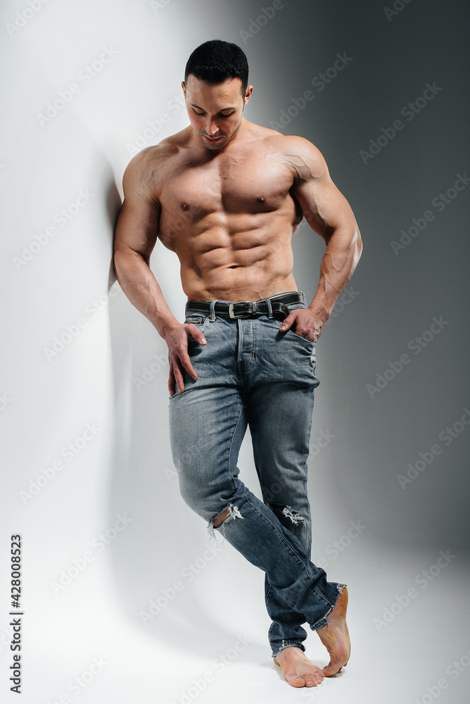 Fototapeta premium A young athlete bodybuilder poses in the studio topless in jeans near the wall. Sport.