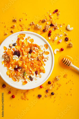 Breakfast concept. Cornflakes, nuts, seeds, berries and honey in a bowl with milk on a bright yellow background. Flakes and nuts scattered on the background. Healthy food. Top view. 