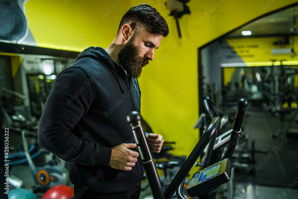 Adult caucasian male athlete training on the treadmill at the gym - back view of man with beard in black hoodie running on the treadmill sport and fitness concept back view copy space