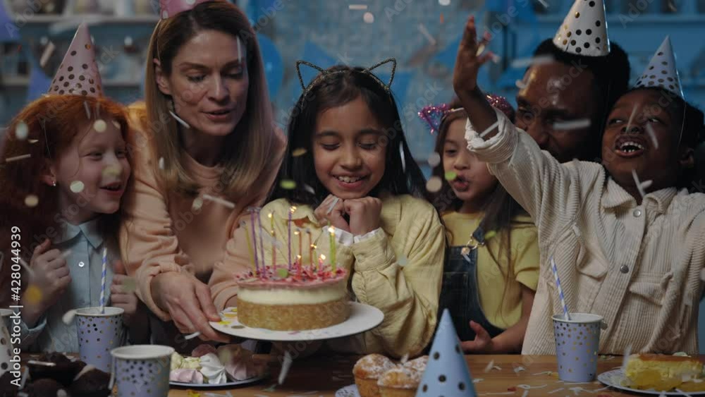 Cute kid with black hair waiting with closed eyes while woman bringing and putting birtday cake in front of her. Girl blowing candles on cake while having party and confetti falling on her.