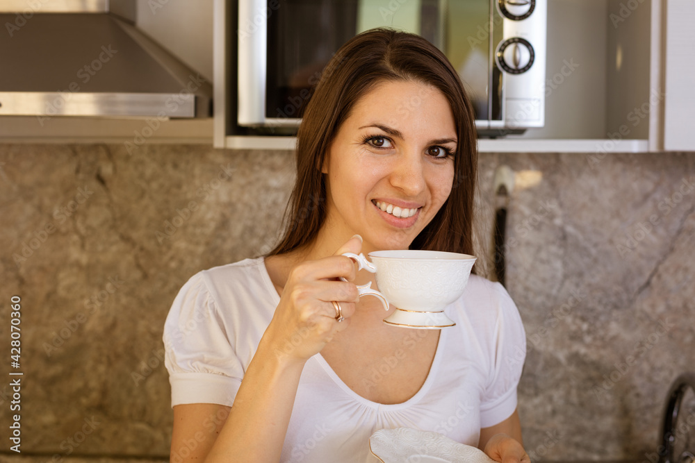 Young beautiful woman in the kitchen sitting at the table with a mug in her hand smiling Young beautiful woman in the kitchen sitting at the table with a mug in her hand smiling