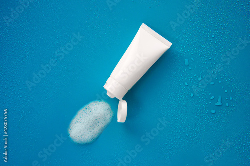 Top view blank label facial cleanser skincare white tube bottle with lid open product squeezed foam soap water texture on plain solid soft blue background with droplets
