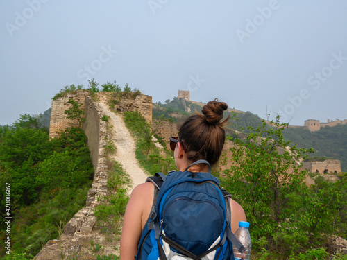 A woman with hiking backpack walking around a unrenewed Gubeikou part of Great Wall of China. The wall is spreading on tops of mountains. Dense forest around it. World wonder. Tradition and history photo