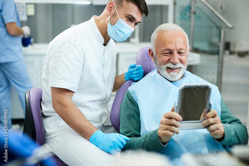 Satisfied senior man looking his teeth in a mirror after dental procedure at dentist's office. photo