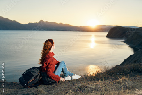 a traveler in a sweater sits on the ground in the mountains near the sea and looks at the sunset