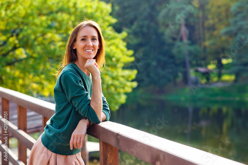 A young, positive, smiling woman stands on a wooden pier, a blurry lake landscape in the background.