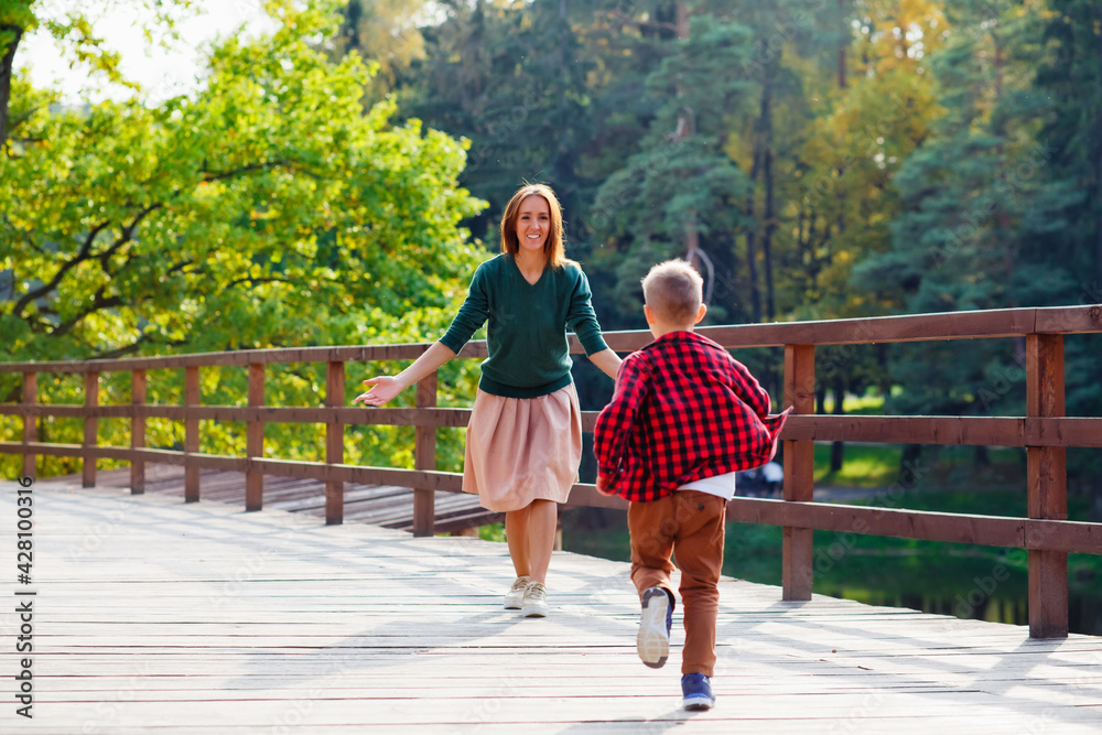 Obraz premium A beautiful woman and a boy on a wooden bridge. Selective focus. A son in a red checked shirt runs to meet his mother.