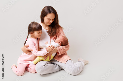 Young beautiful dark haired mom with her 5 years old daughter and newborn baby dressed in casual clothing relaxing and playing together, isolated over white background.
