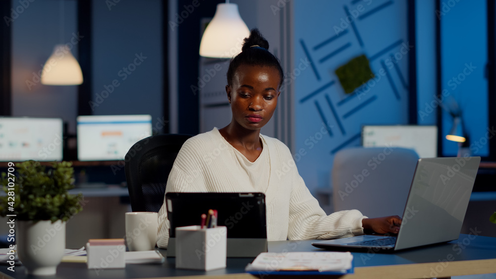 Multitasking black business woman working at laptop and tablet in same time doing overtime in start-up office. Busy african employee analysing financial statistics overworking writing, searching.