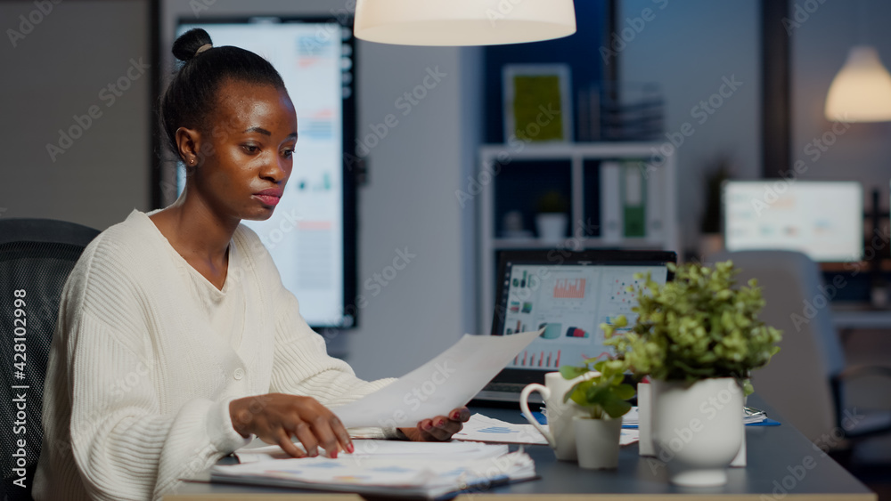 Black manager analysing graphics and financial statistics from documents working overtime in front of laptop sitting in start-up business office. Focused employee respecting deadline of project