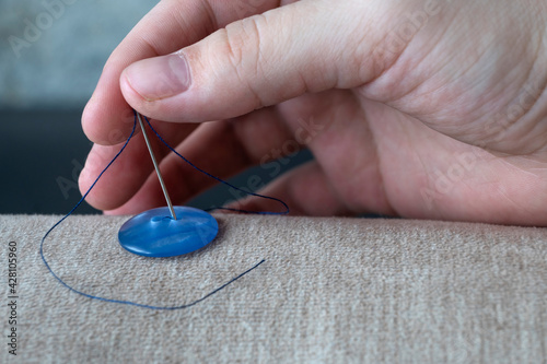 A seamstress's hand sews a blue button with a sewing needle and thread to a rough beige fabric close-up on a gray background. The process of sewing buttons in the sewing industry.